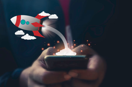 Close up of businessman hand holding mobile phone with rocket flying on screenの写真素材