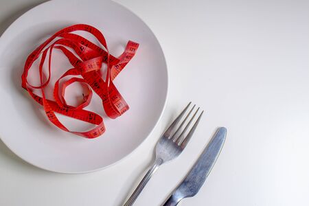Losing weight. A fork and knife next to a white plate with a red measuring tape on a white backgroundの写真素材