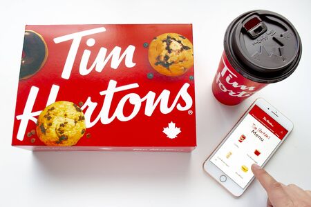 Calgary, Alberta. Canada Jan 27 2020. A person using an iPhone Plus using the Tim Hortons app with a coffee and a box of donuts on a white backgroundのeditorial素材