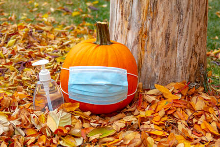 A pumpkin wearing a facemask with a hand sanitizer bottle next to with leaves during fall. Concept halloween and thanksgiving during a pandemic, covid-19, coronavirus.の写真素材