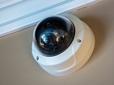 A white dome security camera mounted on a ceiling, with a black lens reflecting the surrounding...