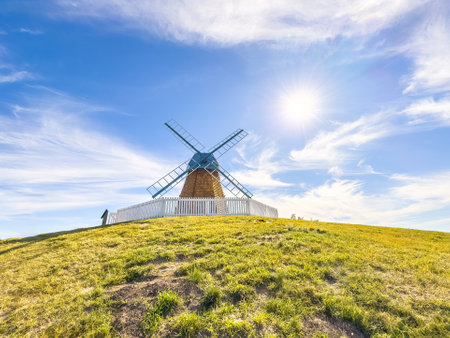 An old windmill with weathered wood and blue sails stands tall on a grassy hill, framed by a white picket fence and a clear blue sky.の写真素材