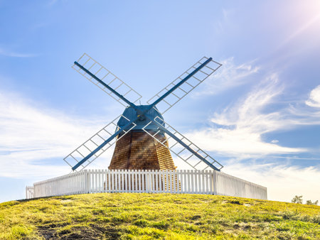 An old windmill with weathered wood and blue sails stands tall on a grassy hill, framed by a white picket fence and a clear blue sky.の写真素材
