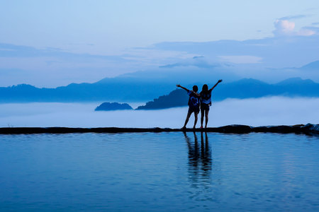 Silhouette rear view of young woman standing near the pool for amazing landscape of blue sky and mountains in morning fog. Happy freedom for success and bliss concept.の写真素材