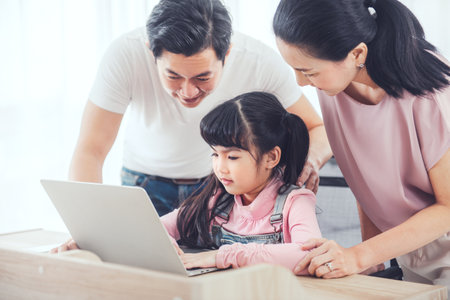 Attractive asian family (mother, father, child daughter) smiling and using laptop computer together while sitting at table in home.の写真素材