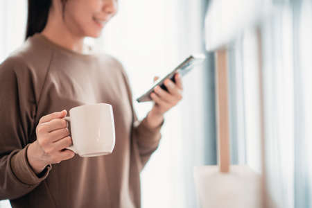 Closeup - Woman hand holding a cup of tea or coffee warm using mobile phone at home, copy space.の写真素材