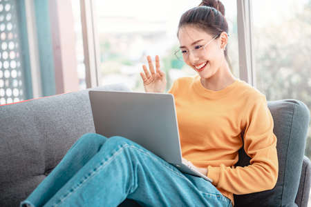 Happy asian woman using videoconferencing with her friend via online app on laptop, waving hand and saying Hiの写真素材