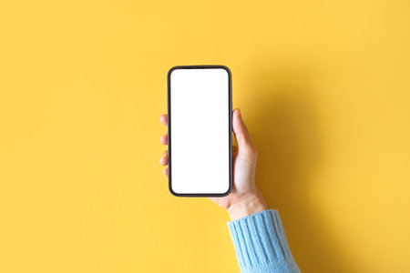 Close up hand holding black smartphone with white screen on yellow background. Mobile phone frameless design concept.の写真素材