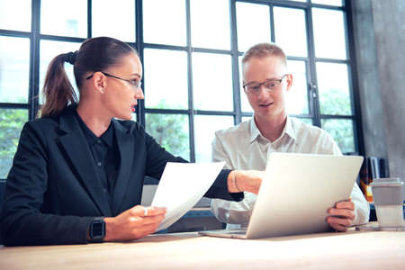 Two diverse business people chatting sitting front laptop in office. Male sharing ideas or startup business plan with female coworker. Business planning.の写真素材