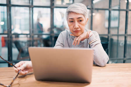 Thoughtful confused mature asian business woman concerned thinking about online problem looking at computer laptop, Worried senior middle aged female reading bad email news.の写真素材