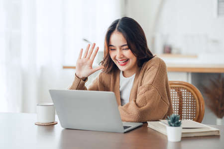 Happy positive young asian woman enjoying online communication at home, Female using wifi while video conferencing with friend, sitting in front of open laptop, smiling and waving hand, saying hiの写真素材