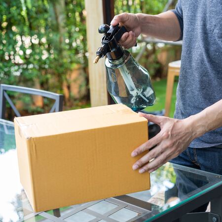Safety delivery service courier during the Coronavirus (COVID-19) pandemic, spraying alcohol disinfectant spray on hands over cardboard box.の写真素材