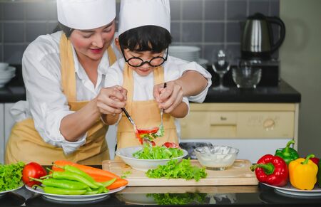Asian woman young mother with son boy cooking salad mom sliced vegetables food son tasting salad dressing vegetable carrots and tomatoes bell peppers happy family cook food enjoyment lifestyle kitchenの写真素材