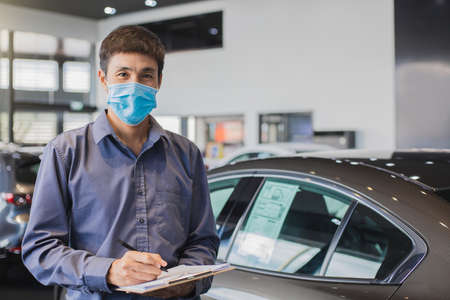 Asian saleman wear surgical mask working in inspector checking writing on clipboard in garage of dealership Wait mechanic In showroom car and insurance new normol covid-19 in showroom dealershipの写真素材