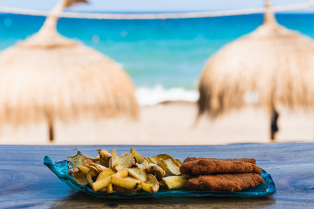 Blue plate of chicken and chips in front of the beach with two umbrellas in the backgroundの写真素材