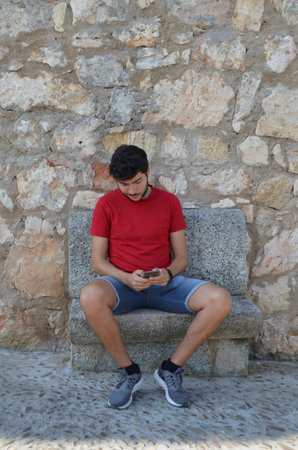 Young Caucasian man in red T-shirt looking at his smartphone sitting on a stone bench looking down and copyspace.の写真素材
