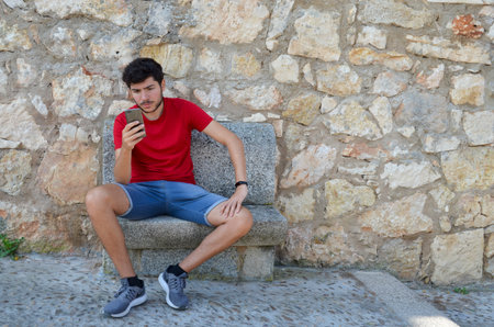 Young Caucasian man in a red T-shirt looking seriously at his smartphone sitting on a stone bench looking straight ahead with copyspace.の写真素材