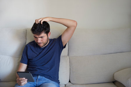 Worried young man scratching his head reading an ebook on the sofaの写真素材