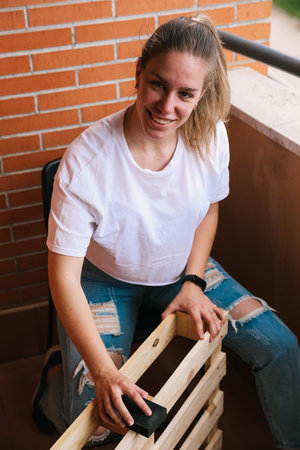 Vertical photo of a smiling blonde woman sanding with hand sandpaper a pallet on the terrace. home renovationの写真素材