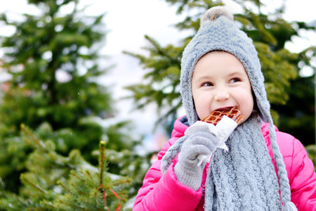 Adorable little girl eating warm waffle on cold winter day near Christmas treeの写真素材