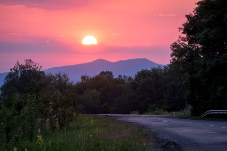 the sun rises against the background of the mountains. Green grass and a road stretching into the distance. Stanitsa Dakhovskaya, Republic of Adygea, Russia.の写真素材