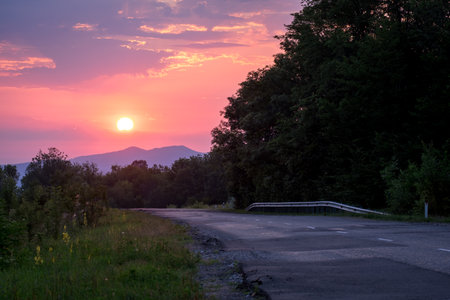 Country road at sunrise. The sun rises against the background of the mountains. Green grass and a road stretching into the distance.の写真素材
