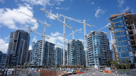 A vibrant urban construction site showcasing modern buildings with glass facades, multiple tower cranes, and a clear blue sky, emphasizing growth and development.の素材