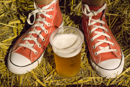 Female legs wearing red canvas sneakers. Plastic cup of lager beer between them. Ground strewn with straw. Night outdoor scene on the music festivalの写真素材