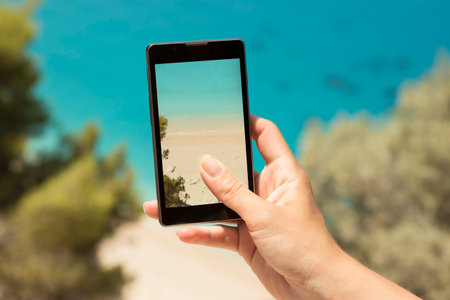 Female hand taking a photo of a beautiful sandy beach by using the smartphone. Pristine landscape.  の写真素材