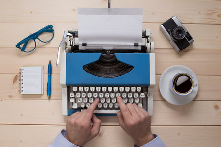Journalist working on typewriter at his desk. No recognizable person. Top down view point. Along with typewriter there are eyeglasses, notebook, ball pen, cup of coffee and retro camera on the table     の写真素材