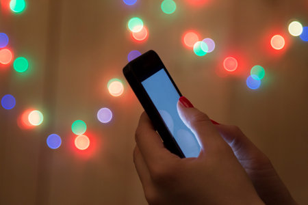 Female hand holding cellphone and colorful bokeh lights in the background.の写真素材
