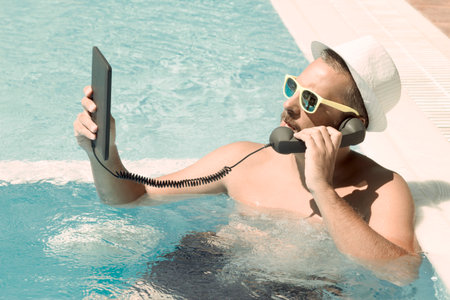 Young man having an internet call in a swimming pool. He is using digital tablet device and retro styled handset.の写真素材