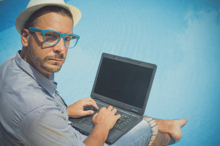 Portrait of a man using laptop by the swimming pool. He is wearing hat and blue eyeglasses. Freelancer concept. の写真素材