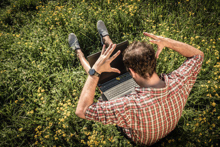 Guy sitting on grass with laptop on his knees. He is in stress as something went wrong on his computer. の写真素材