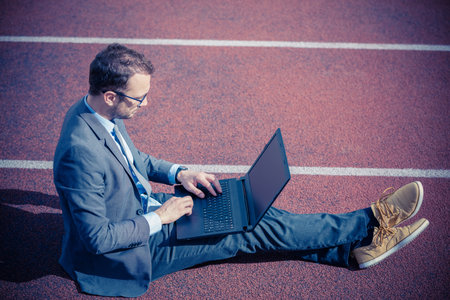 Businessman in suit sitting on jogging track and using laptop. Business race and success concept.の写真素材