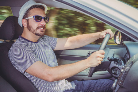 Side point of view of a young man wearing hat, sunglasses and stripped shirt driving a car. Vacation...