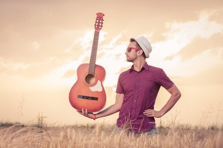 Man with hat and sunglasses holding acoustic guitar in wheat field with sunset and clouds in the...