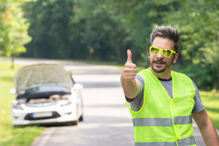 Young modern tourist man in reflective vest standing on the road and showing thumbs up. Car with opened hood in the background. Road assistance and safety measures concept.の写真素材
