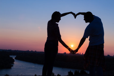 Silhouette of young couple making heart shape with hands on hill above the city at sunset.  の写真素材