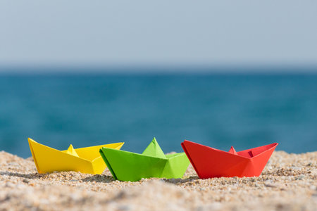 Close up of paper boats in different colors on sandy beach. Ocean in the background. Summer vacation and travel concepts.の写真素材