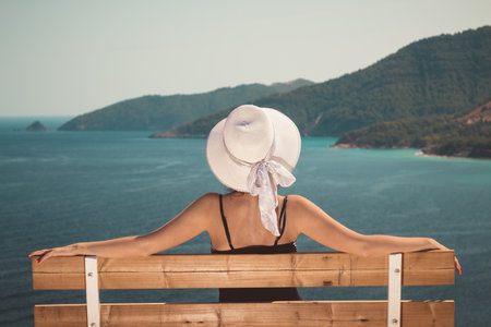 Rear view of stylish female sitting on bench and enjoying the sea view. Travel and vacation concepts.の写真素材
