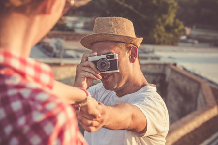 Young trendy guy taking photos of his girlfriend with retro vintage camera and holding her hand in nature. Travel, vacation and love concepts.の写真素材