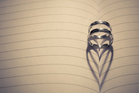 Wedding rings placed at the middle of the blank notebook. Back light forms heart shape shadow. Space for copy. Love and marriage concepts. の写真素材