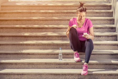 Pretty sportswoman resting on staircase outdoors after fitness training and using smartphone.  の写真素材