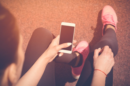 Female jogger sitting on bench next to the jogging track and using smartphone with blank screen. Preparing for the morning workout. Top down point of view. Technology, sport and fitness concepts.の写真素材