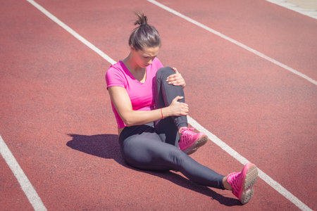 Female sitting on running track and holding her injured leg. Accident during jogging     