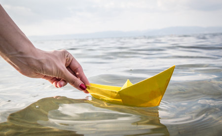 Close up of female hand runs yellow paper ship in sea. Travel and summer vacation conceptsの写真素材
