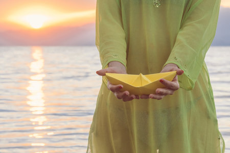 Close up to female hands holding yellow paper boat. Sunset over sea in the backgroundの写真素材