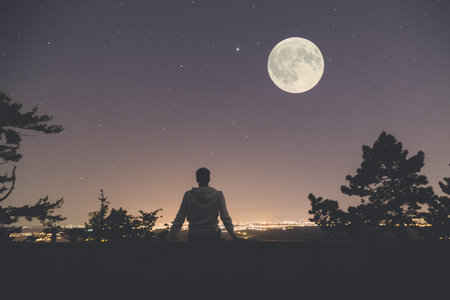 Young man sitting on the wall at night. City lights, moon and stars in the background. の写真素材