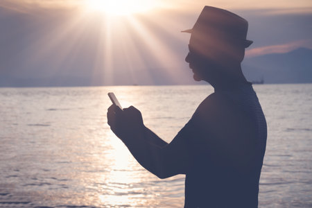 Side view of man in hat using and looking at mobile phone during sunset over the oceanの写真素材